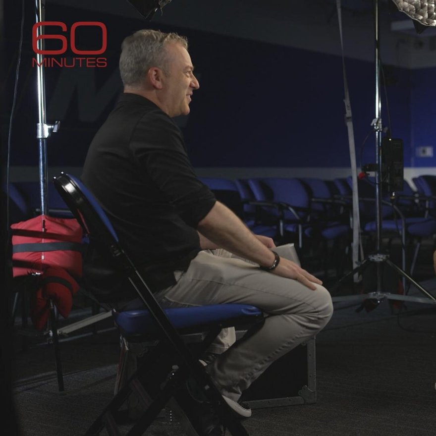 A single lone football sitting in an empty news anchor's chair behind a desk with a ticking stopwatch on it, studio lights, no text.