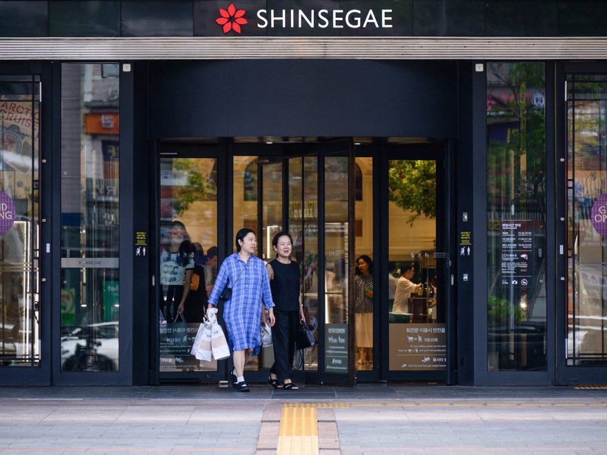 A single glossy luxury handbag sitting alone on a department store display pedestal, a faint green stock chart reflected in the glass behind it.
